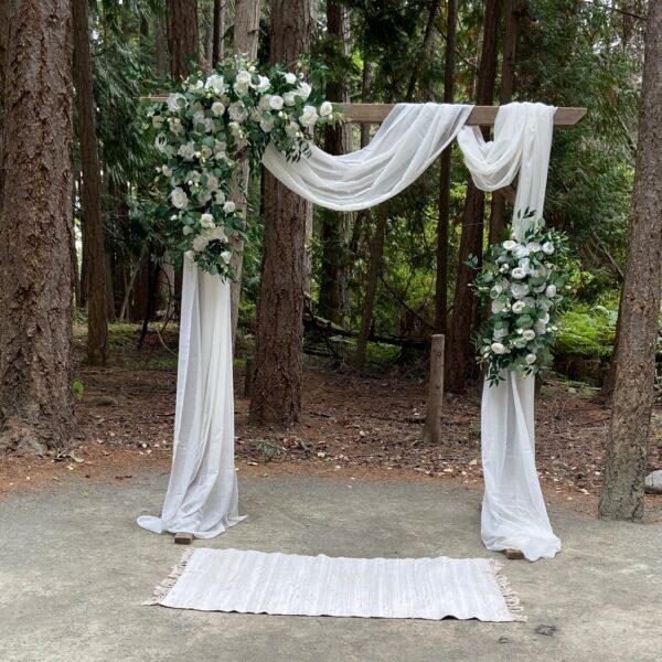 A white arch with greenery and drapes in the middle of a forest.