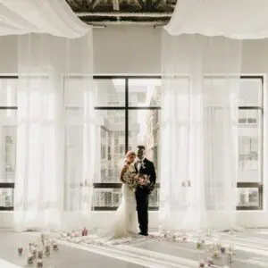 A bride and groom standing in front of windows.